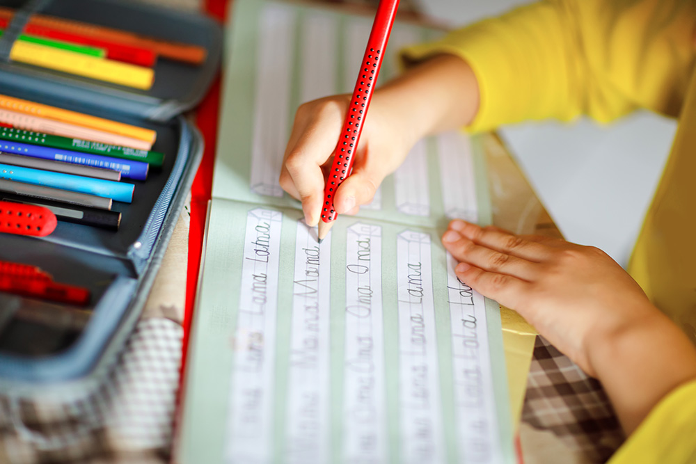 Ein Kind schreibt mit einem Bleistift in ein Übungsheft das Wort Mama in Druckbuchstaben.